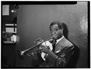 Image of Louis Armstrong leaning against a wall while playing the cornet with eyes wide open.